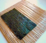 Расстилаем на столе макису, и оборачиваем ее пластиковой пищевой пленкой. Берем лист водорослей нори, разрезаем те пополам, и укладываем параллельно пластинкам бамбуковой циновки, блестящей стороной вниз.