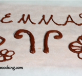 Молочный шоколад растапливаем на водяной бане. Заворачиваем разделочную доску пергаментной бумагой и с помощью кондитерского шприца топленым шоколадом рисуем массивные рожки для бабочки. Из остального шоколада делаем украшения или имя именинника. Ставим на час шоколад в холодильник, чтобы украшение полностью застыло.	