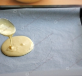 Сначала готовим коржи. Сгущенку и яйца выложите в большую миску и хорошенько перемешайте венчиком до однородного состояния. Просейте в миску муку и разрыхлитель, снова тщательно перемешайте, а затем дайте постоять тесту минут 15, чтобы коржи получились пышными.
Разогрейте духовку. На листе бумаги для выпечки нарисуйте круг, диаметром около 18-20 см. Бумагу с кругом положите на противень и налейте в круг 2 полные столовые ложки теста, разровняйте.